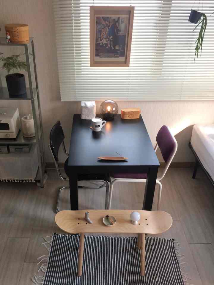 白い壁とブラインドがある狭いダイニングルーム、黒いダイニングテーブルと木目調のスツール、2つのダイニングチェアが配置されたミッドセンチュリーモダン風の空間