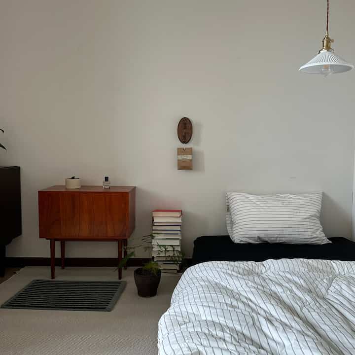 白とベージュの色調の寝室、ストライプ柄の寝具と木製のドレッサーが配置されたシンプルでモダンな雰囲気のあるインテリア