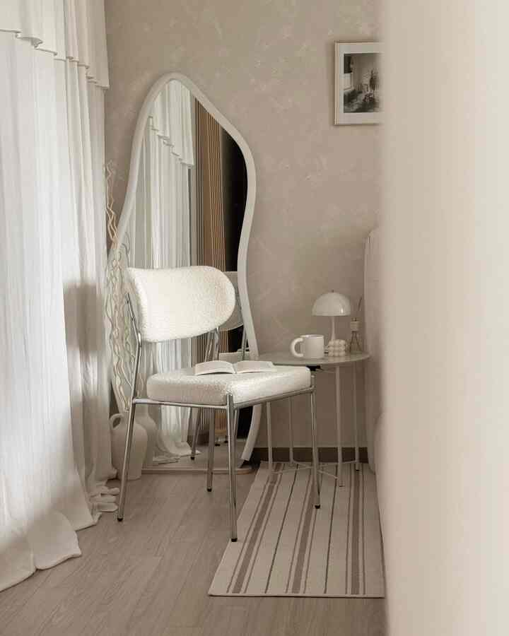 柔らかいベージュとホワイトの色調の小さなコーナースペース、ミニマルなアームチェアとサイドテーブル、大きな不規則形の鏡が配置された雰囲気あるインテリア
