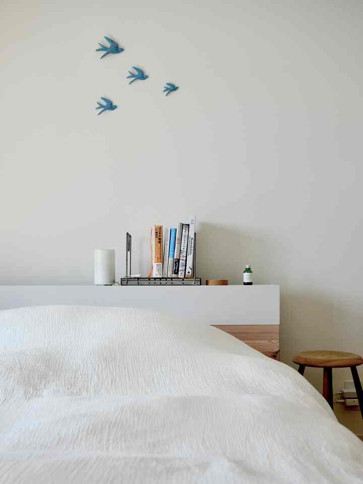 白と木目調の調和がある寝室、シンプルな無印良品の寝具と小さなスツール、本棚、壁に鳥の飾りが配置された雰囲気のあるインテリア