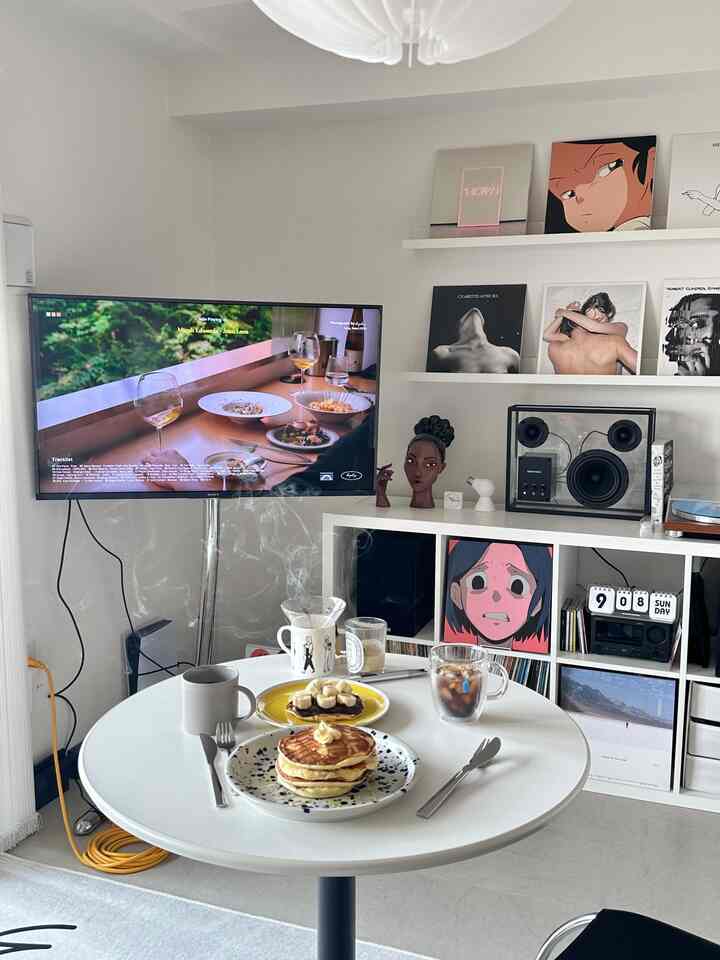 白を基調としたリビング、丸テーブルにパンケーキとコーヒーが置かれ、壁の棚とテレビ台が配置されたモダンなインテリア