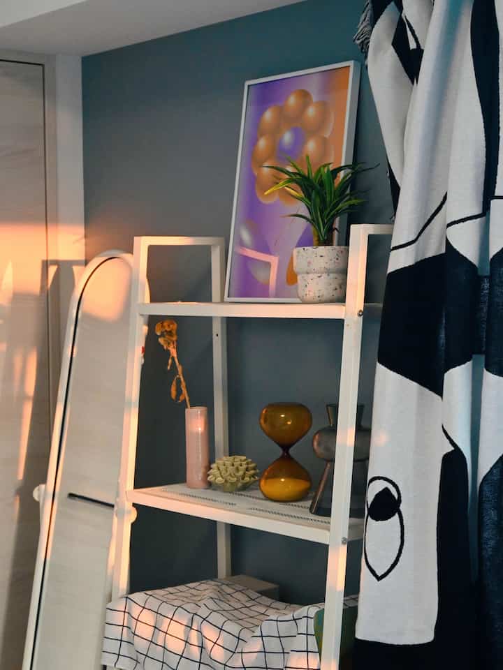 A compact room featuring a white metal shelf with a poster and plant, alongside black patterned curtains, creating a natural and modern atmosphere