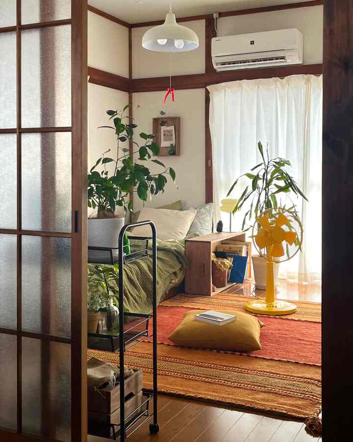 ブラウンの木目調とナチュラルなグリーン植物が調和した暖かみのある寝室、オレンジのラグと黄色の扇風機がアクセントのあるインテリア
