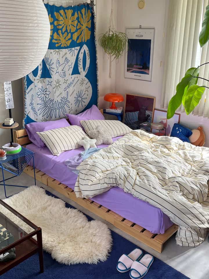 ラベンダーとブルーの色調の寝室、木製のパレットベッドと多様なクッションが配置された居心地の良いボヘミアンスタイルのインテリア