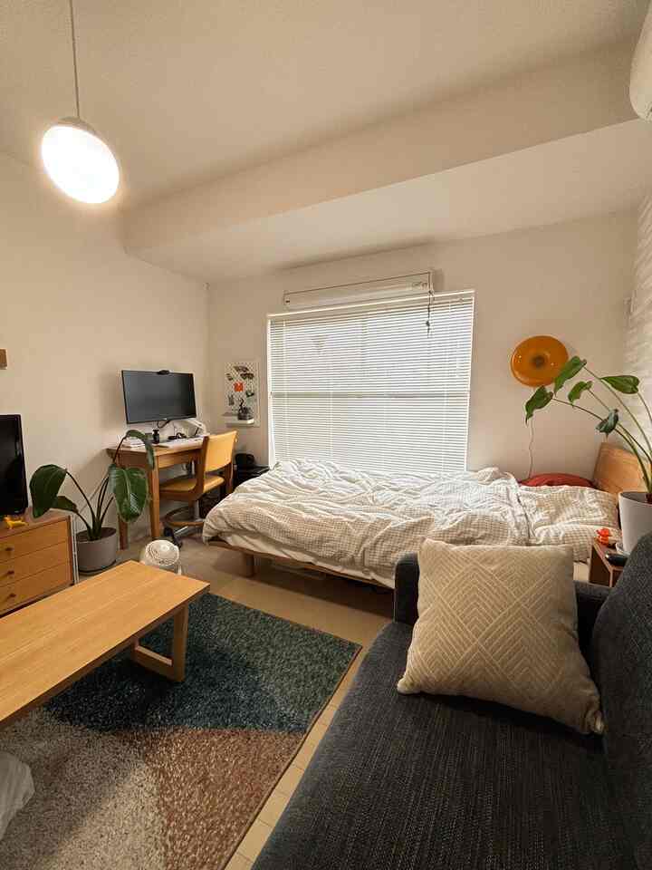 Natural tone 1K studio apartment featuring a bed by the window, wooden desk with monitor, and dark sofa in a cozy living area