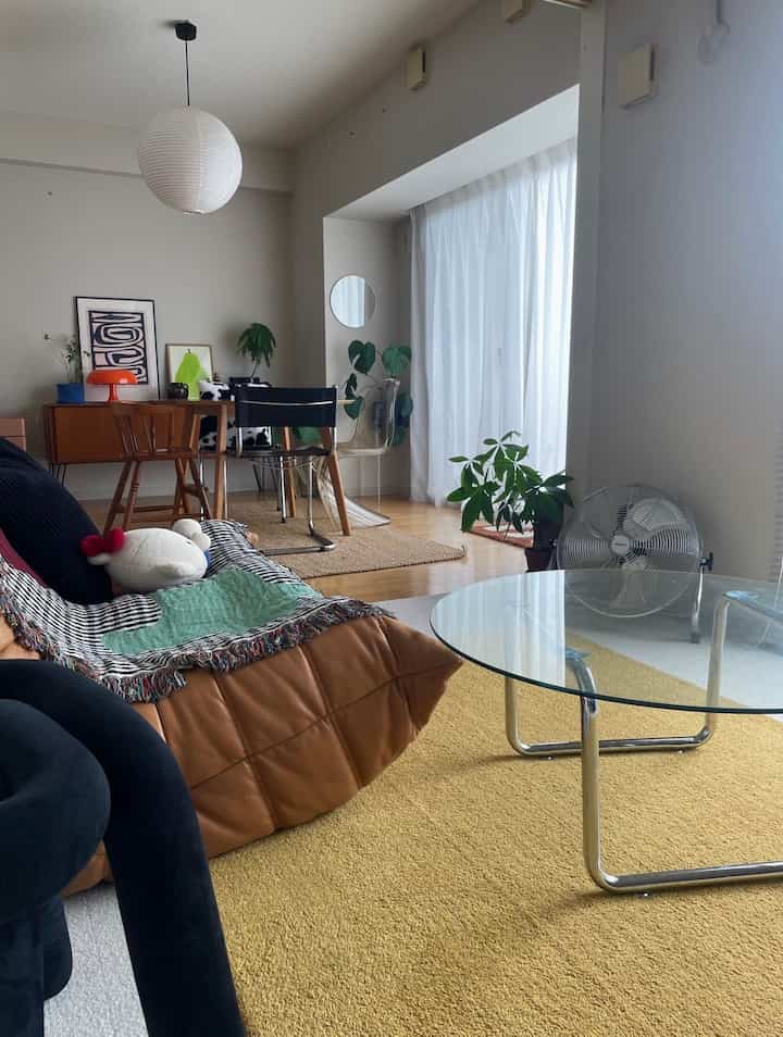 A natural-toned living room with white and brown hues featuring a brown sofa with a colorful throw and round glass coffee table