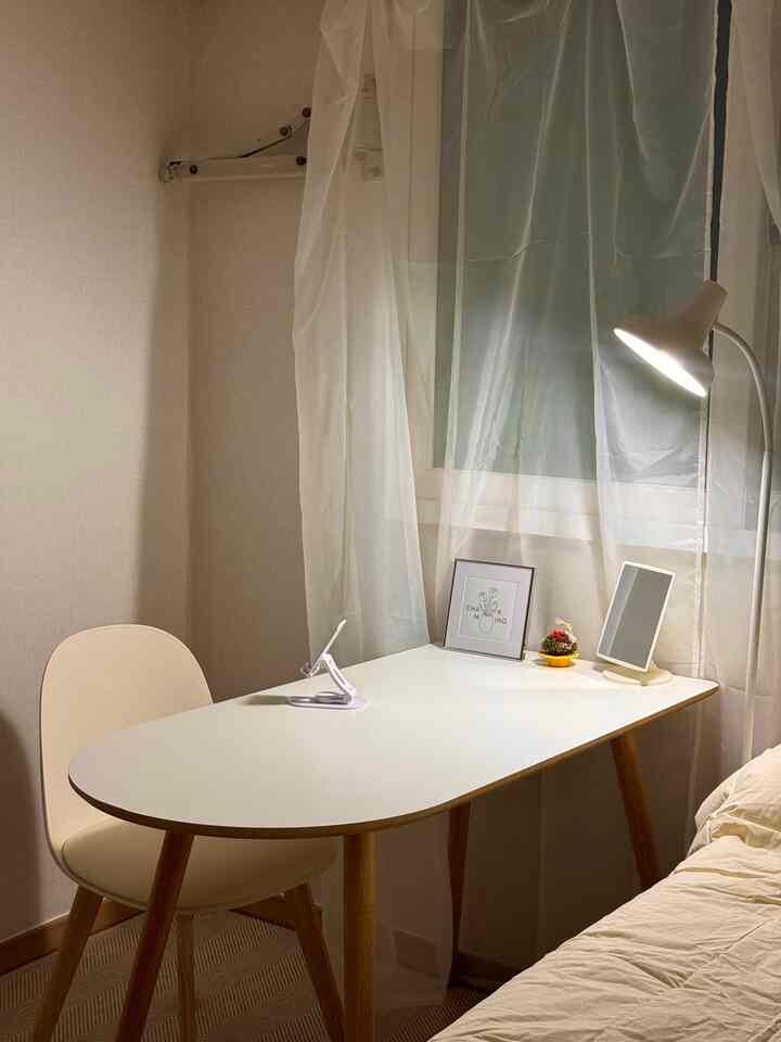 木目調の脚が特徴的な白いテーブルとベージュのベッドがあるコンパクトなワンルーム、フロアランプで照らされた空間