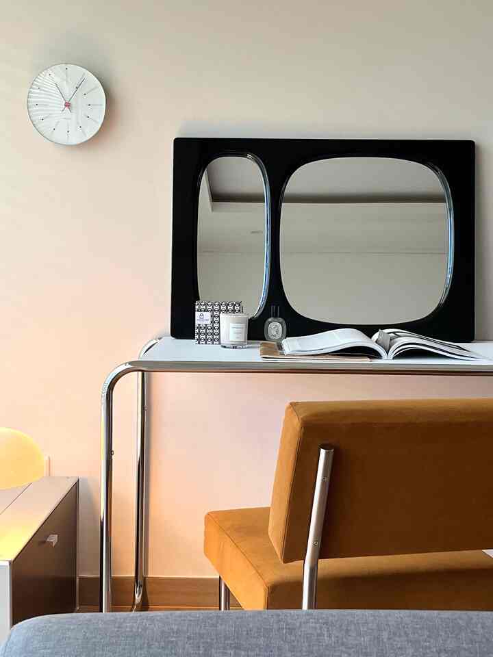 柔らかなベージュの壁とクローム脚の白い化粧台、黒いフレームの鏡、茶色のベルベットチェアが配置されたミッドセンチュリーモダンな空間