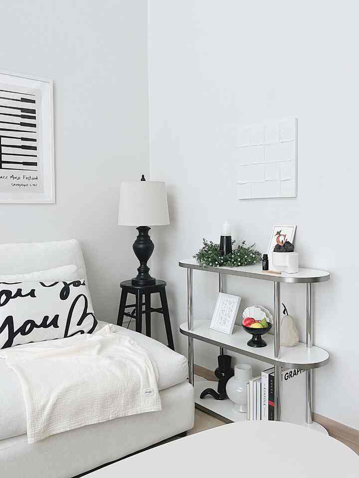 A white and black toned living room photo zone featuring a white sofa, 3-tier shelf, elegant lamp, and decorative objects in a clean, modern space