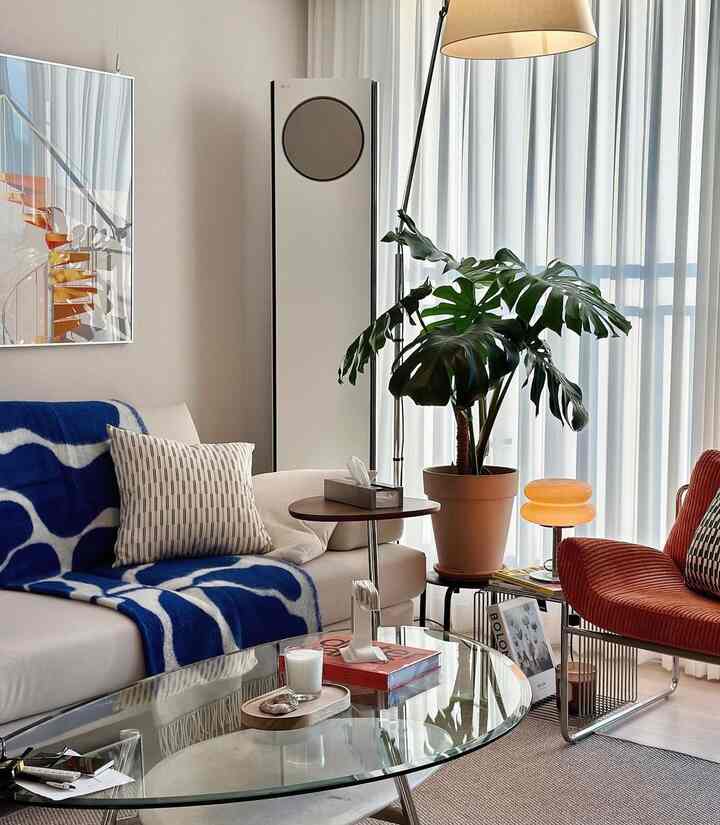 ベージュとオレンジの色調のリビング、透明なガラステーブルとオレンジのアームチェア、大きなモンステラの植物が配置された温かみのある雰囲気