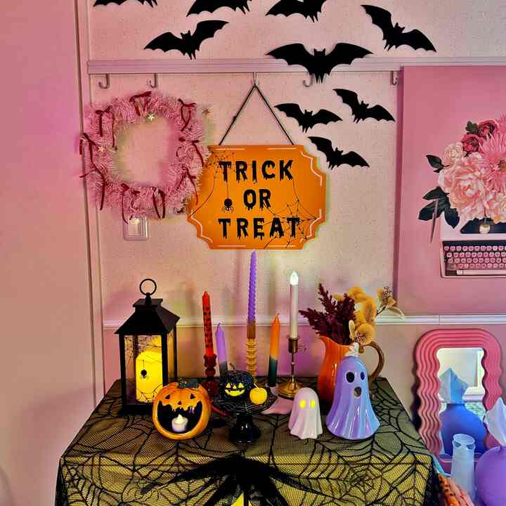 ピンクの壁と黒のクモの巣模様のテーブルクロスがあるハロウィンの飾りつけ空間、黄色い「TRICK OR TREAT」サインや様々なキャンドル、ゴーストのオーナメントが配置された雰囲気のあるインテリア