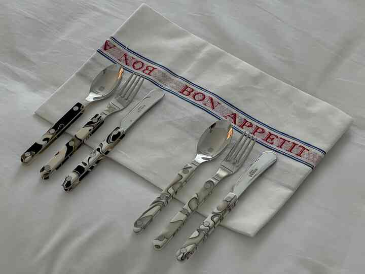 白い背景に柄のあるサーブルのデザートカトラリーとボナペティのキッチンクロスが配置された、清潔感のあるテーブルウェア
