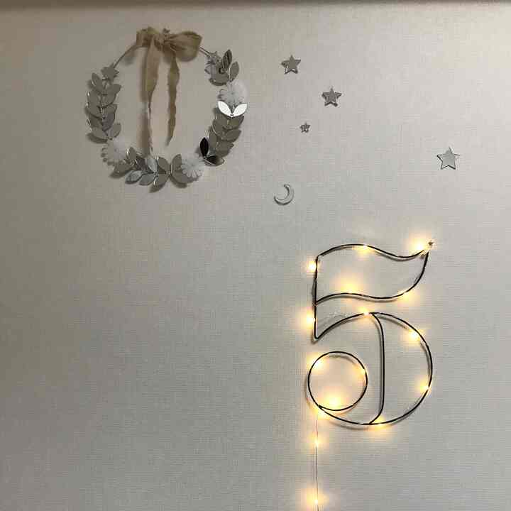 White-toned kids' room wall featuring star and moon stickers, illuminated number '5' objet, and a mirror leaf wreath in a minimal setup