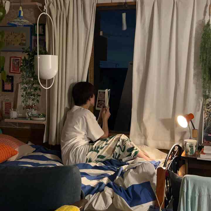 A cozy modern bedroom in beige and blue tones featuring a bed with striped bedding and a person reading by the window