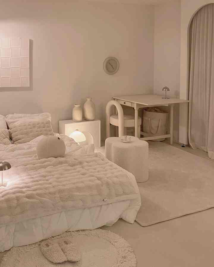 アイボリーとナチュラルトーンの寝室、マットレスの上にブランケットとスツール、右側にテーブルと照明が配置された温かな印象の空間