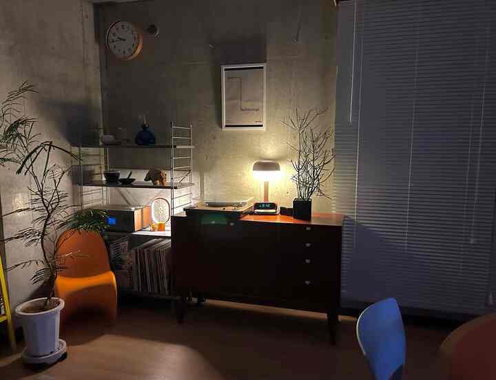 グレーのコンクリート壁を背景にしたナチュラルモダンなリビング、オレンジのパントンチェアと木製キャビネット、フロアランプと植物が配置された印象的な空間