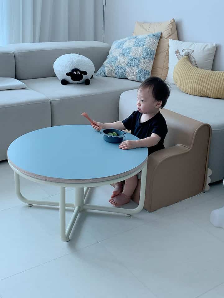 ベージュとスカイブルーの色調のリビング、赤ちゃん用チェアと丸テーブルが配置されたシンプルで雰囲気のある空間