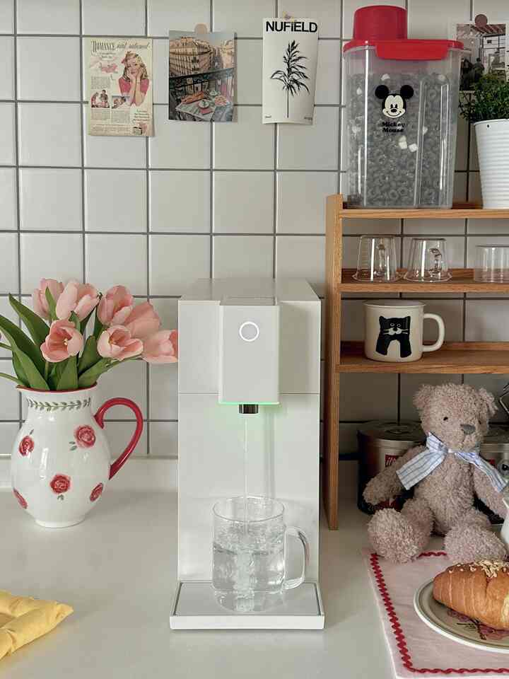 白と木目調が調和したキッチン、中央に浄水器があり、棚には猫のマグカップとテディベアが置かれた雰囲気のあるインテリア