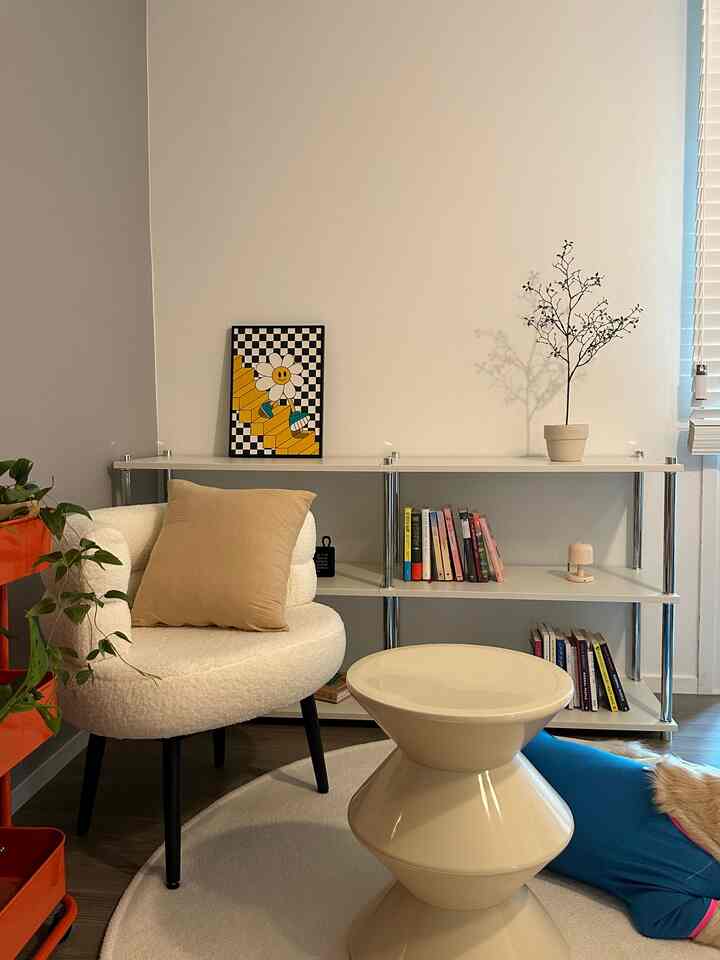 アイボリーとベージュのトーンの書斎、丸いスツールとブークレのアームチェア、本棚に小物が置かれたシンプルでモダンなインテリア