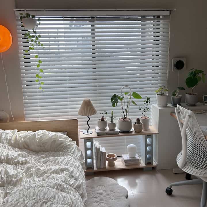 明るいホワイトと木目調の寝室、窓辺の棚に植物と本が配置された雰囲気のあるインテリア