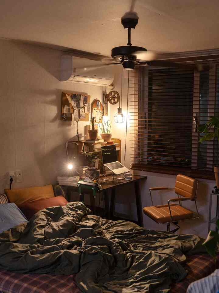 ウッドトーンとナチュラルな植物が調和した、一人暮らし向けの寝室とホームオフィスのデスクゾーンが温かい照明の下に配置された居心地の良い空間