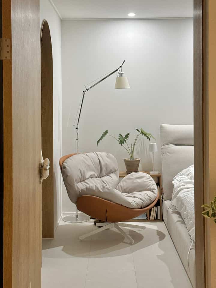 ベージュとブラウンの落ち着いた寝室、回転式の一人掛けソファとフロアランプ、サイドテーブルが配置された雰囲気のあるインテリア