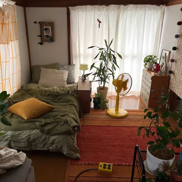 緑の寝具と複数の植物が配置された狭い寝室、黄色の扇風機とラグがアクセントのナチュラルな雰囲気のインテリア