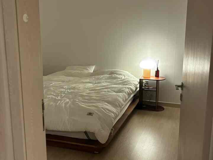A white and wood-tone bedroom featuring a low wooden bed frame and a nightstand with a table lamp, creating a cozy atmosphere