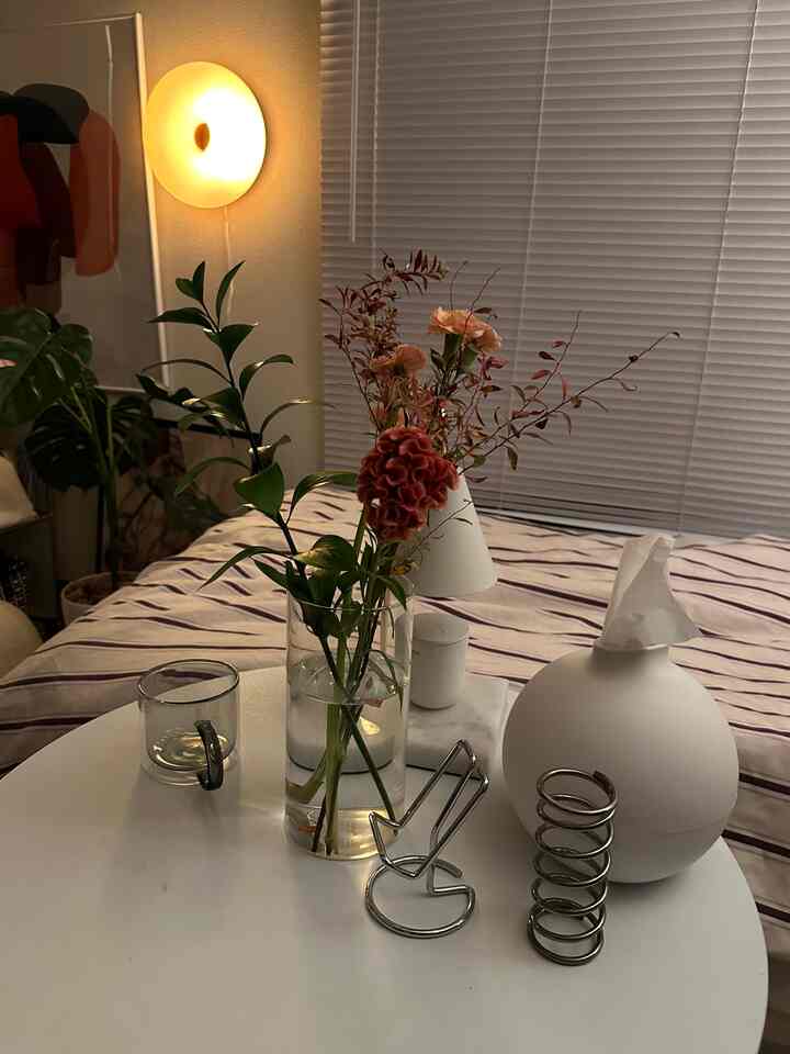 暖かな照明と植物があるナチュラルな色調の寝室、白いブラインドと花瓶が置かれたテーブルが配置された居心地の良い空間