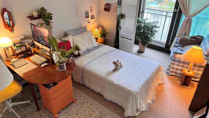 暖かい黄色の照明と木目調の色合いがある寝室兼作業スペース、窓際に植物とチェック柄のアームチェアが配置された雰囲気のある空間