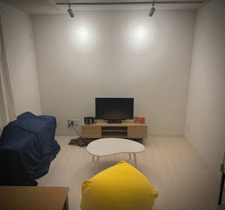 白い壁と木目調の家具が配置されたシンプルなリビング、黄色いビーズクッションとコーヒーテーブルが印象的な空間