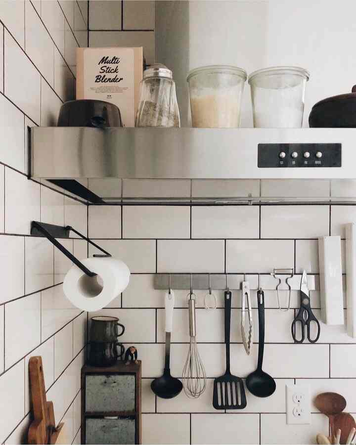 白いタイル壁とステンレスフードのあるキッチン、黒い調理器具と茶色の木製道具が整然と配置された清潔感のある空間