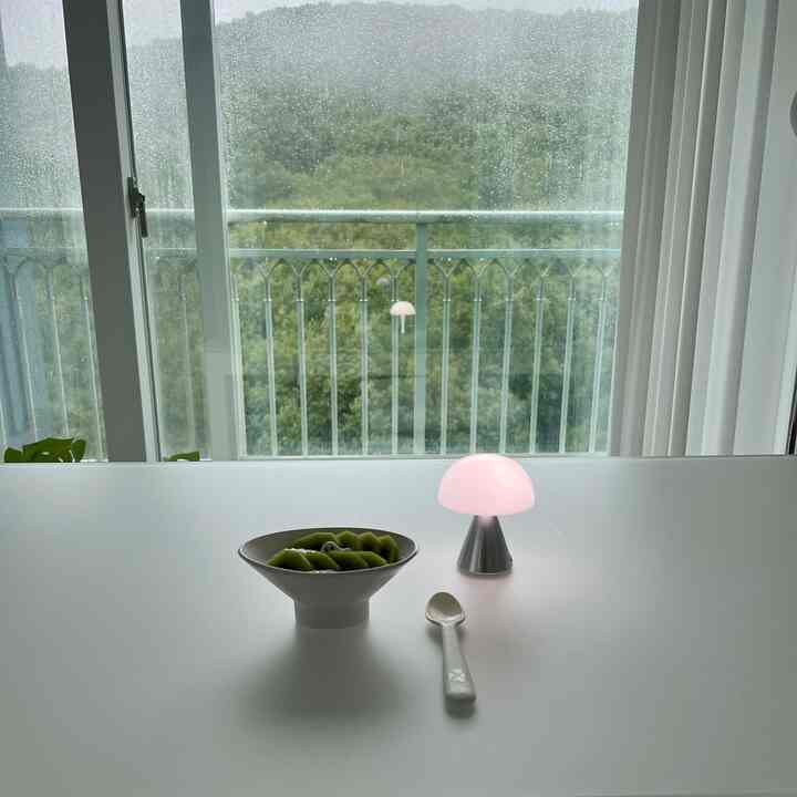雨の降る窓越しに自然の景色が見え、白いテーブルの上にボウルとLEDマッシュルームランプが置かれたモダンなダイニングインテリア