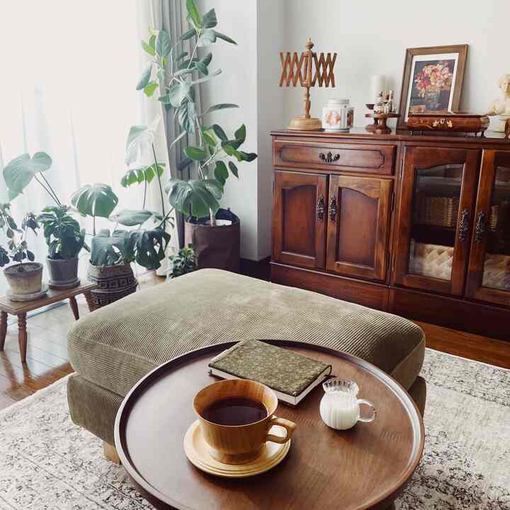 温かみのある木目調と自然の植物が調和するヴィンテージ風リビング、オットマンとサイドテーブルが配置された居心地の良い空間