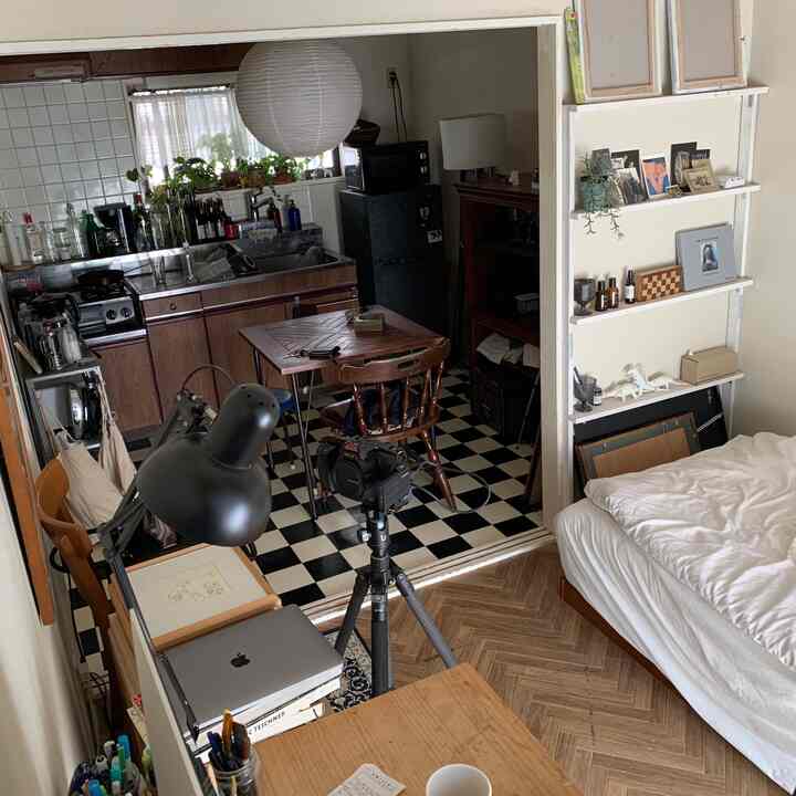White-walled 1K apartment with wood-toned furniture, featuring a bedroom and kitchen connected in a cozy, cluttered single household space.
