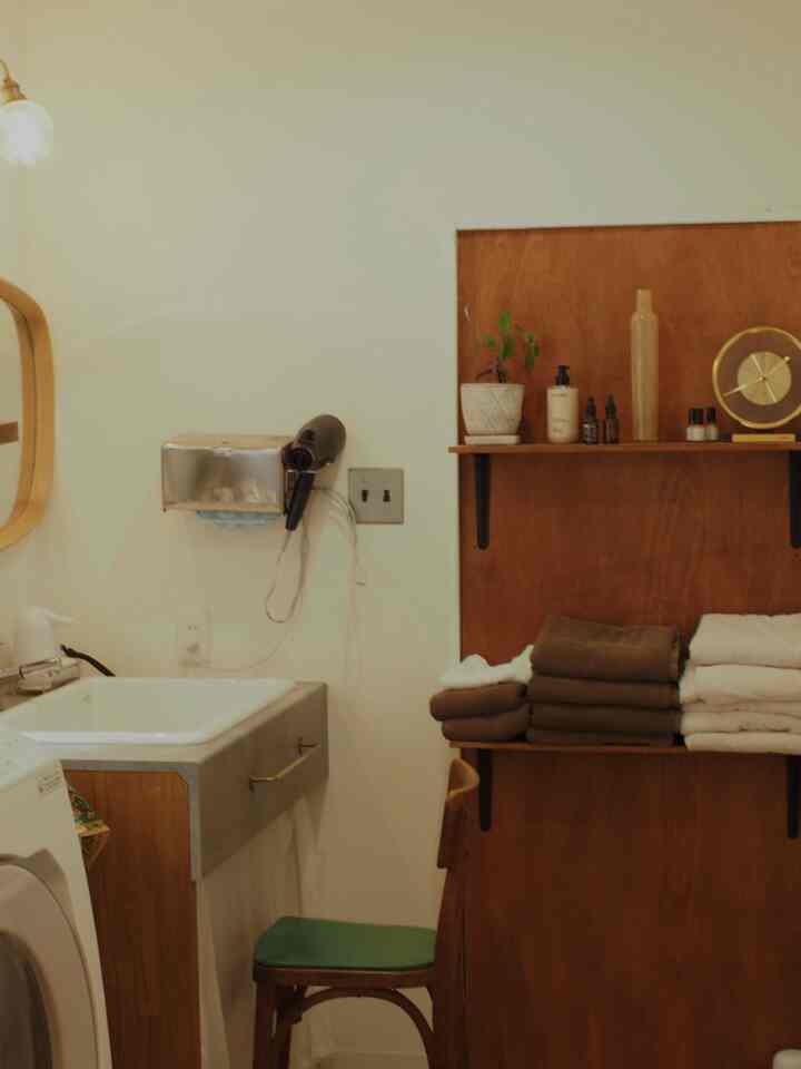 白い壁と茶色の棚があるバスルーム、洗面台の周りにタオルと椅子が配置された温かみのある空間