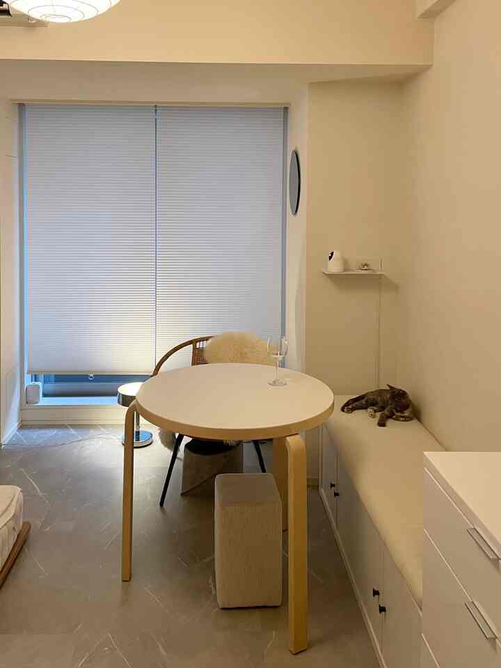 Beige-toned compact room featuring an Artek round table, blinds, and a cozy cat lounging on a bench in a natural setting.