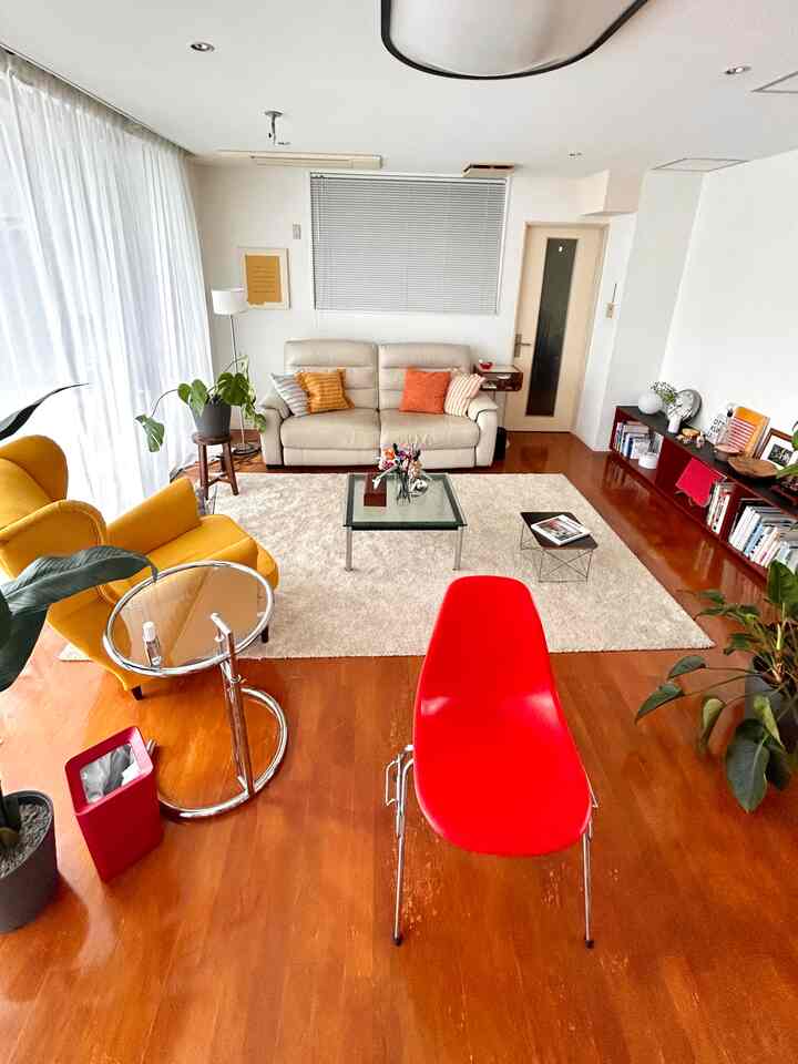 白い壁と木目調の床のリビング、ベージュのソファと黄色いアームチェア、赤い椅子が配置された明るく自然な雰囲気のインテリア