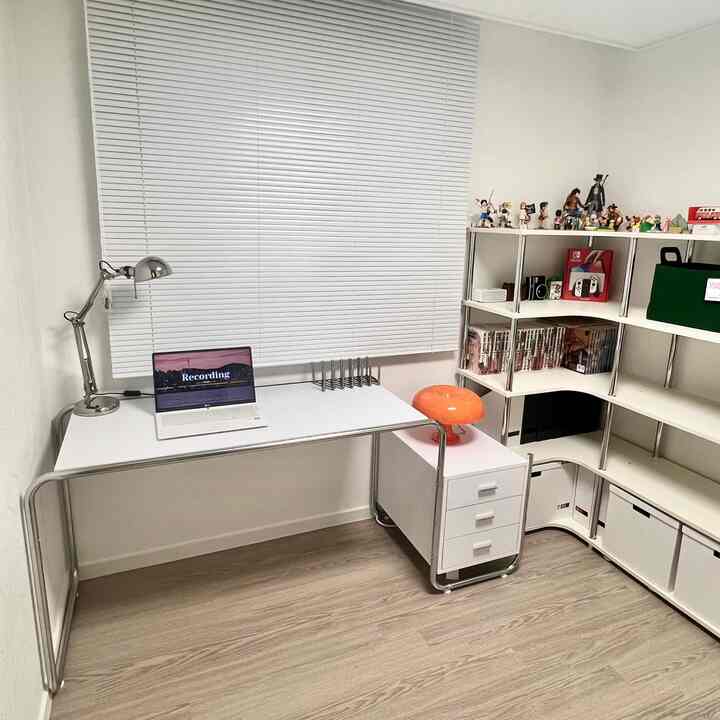 ホワイトとクローム調の小さな書斎、ホワイトのデスクとL字型の棚にフィギュアや各種アイテムが配置された、すっきりとした趣味部屋のインテリア