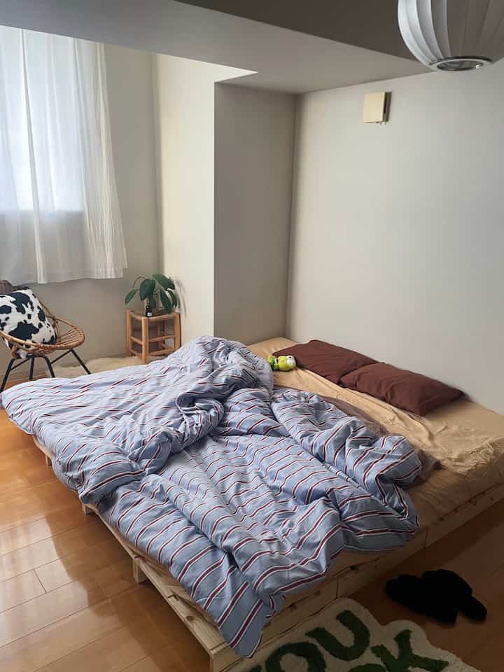 ナチュラルな木目調とパステルブルーの寝具が調和するコンパクトなワンルーム寝室。フロアベッドとラタン製アームチェアが配置された空間