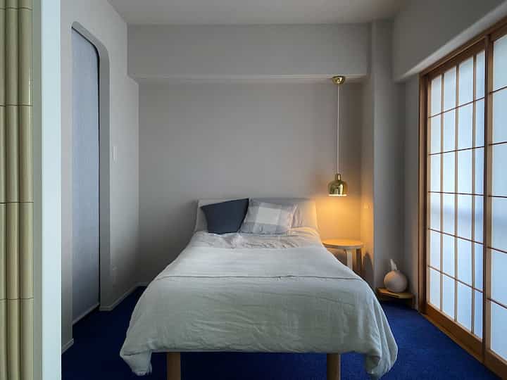 ブルーのカーペットと白い寝具が配されたミニマルな寝室、ゴールドのペンダントライトが雰囲気を演出する空間