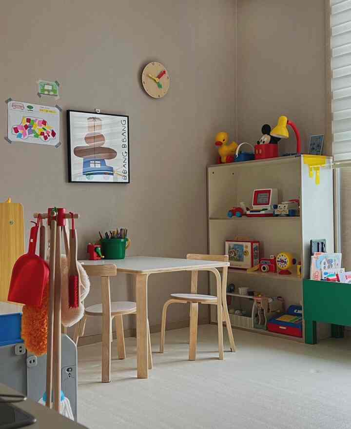 ベージュの壁と床の子供部屋、木製の机と椅子2脚、おもちゃが並ぶ収納棚が配置された雰囲気のある遊び空間