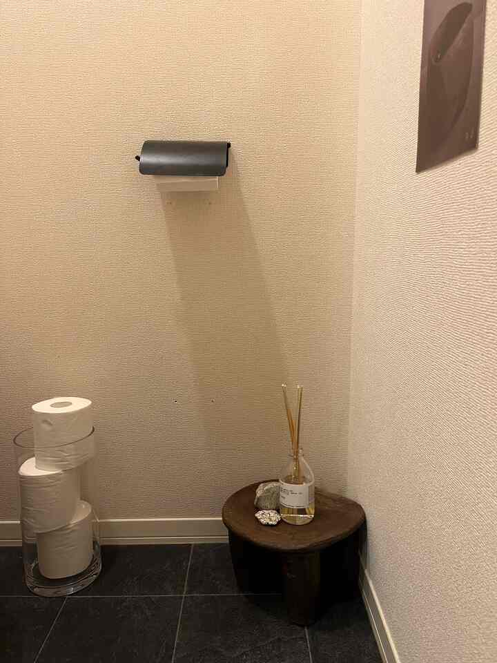 ベージュ調の壁のトイレ、透明なガラス花器に置かれたトイレットペーパー、小さな木製スツールとリードディフューザーが配置された清潔なインテリア