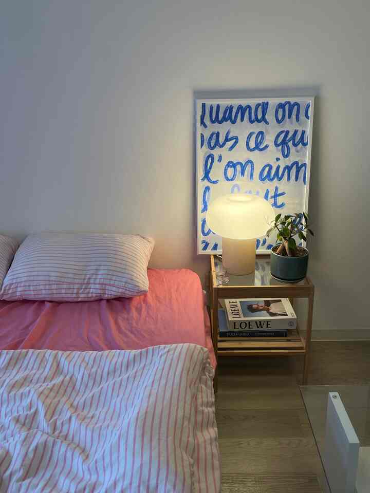 A cozy studio bedroom with pink bedding and a blue poster, featuring a beige table lamp and wooden nightstand