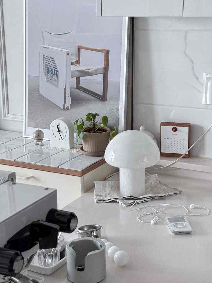 白を基調としたキッチンの一角にヴィンテージ風のガラステーブルランプと植物、カレンダーが配された雰囲気のある空間