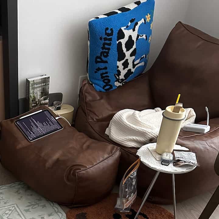 Cozy living room featuring brown bean bag sofa with a blue cushion, small side table with tumbler, and white walls, suited for reading and home cafe ambiance