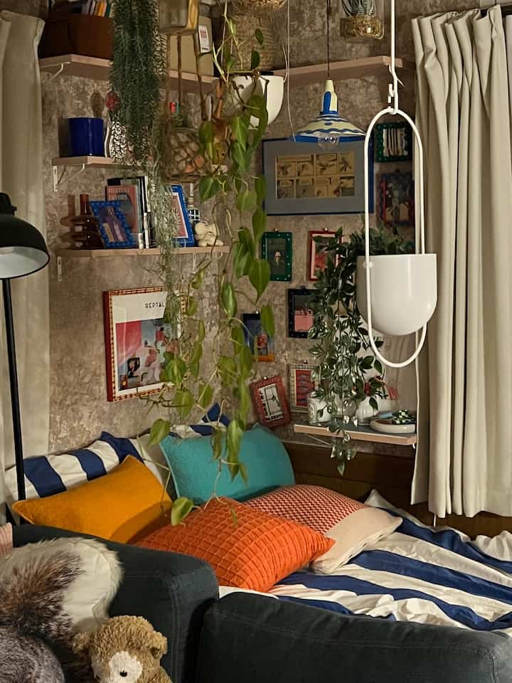 青と白のストライプ寝具とオレンジのクッションが並ぶ寝室、植物と間接照明が配された雰囲気のあるカフェ風インテリア