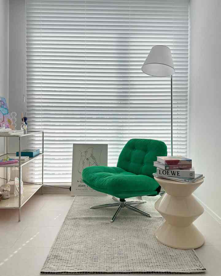 白い壁とブラインドを背景にした子供部屋、緑の回転アームチェアとフロアランプ、ストライプのラグが配置された雰囲気のある読書スペース