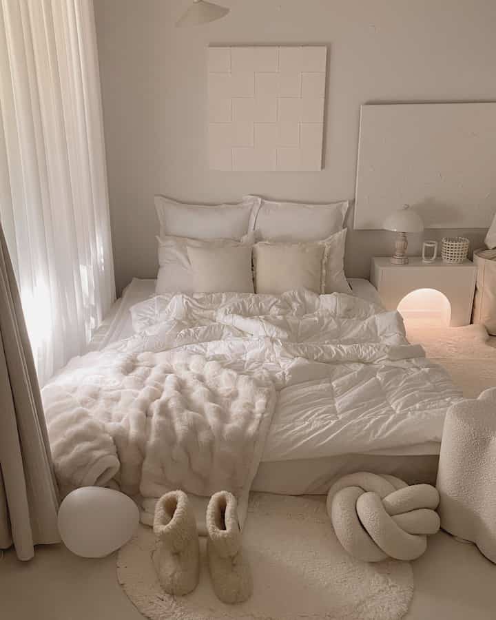 明るい白とクリーム色調の寝室、ヘッドレスベッドと暖かなブランケット、枕が整然と並ぶ居心地の良い空間