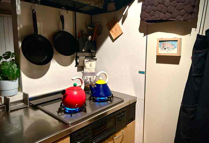 モダンなキッチン、赤と青のやかんがコンロの上に置かれ、壁にフライパンや調理器具が整然と掛けられた一人暮らしのキッチン